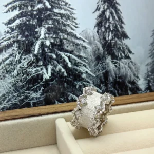 The image features a stunning Sterling Silver Lace Edge Ring named Northern Elegance. The ring is displayed in an open jewelry box, positioned against a backdrop of snow-covered pine trees, adding a festive touch to the scene. The Lace Edge Ring showcases an intricate lace pattern along its edges, which enhances its elegant and sophisticated appearance. The reflective sterling silver material shines beautifully, capturing the light in a way that highlights its detailed craftsmanship. This Sterling Silver Ring makes an ideal Christmas gift, combining timeless elegance with a festive presentation.