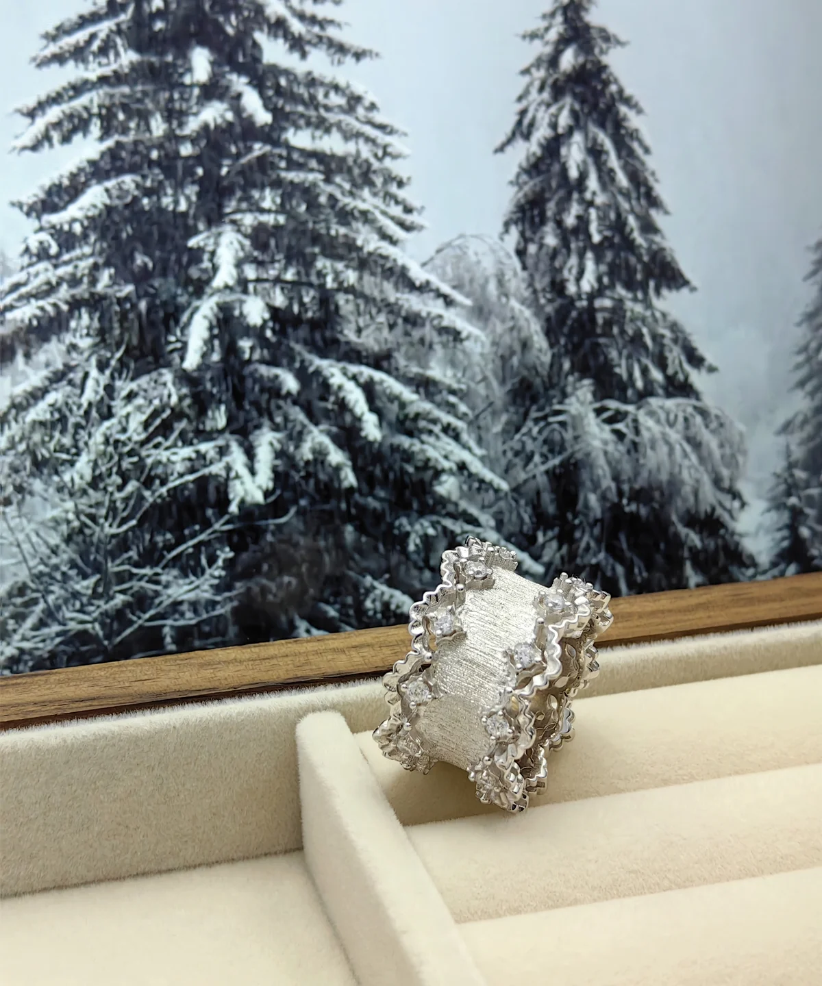 The image features a stunning Sterling Silver Lace Edge Ring named Northern Elegance. The ring is displayed in an open jewelry box, positioned against a backdrop of snow-covered pine trees, adding a festive touch to the scene. The Lace Edge Ring showcases an intricate lace pattern along its edges, which enhances its elegant and sophisticated appearance. The reflective sterling silver material shines beautifully, capturing the light in a way that highlights its detailed craftsmanship. This Sterling Silver Ring makes an ideal Christmas gift, combining timeless elegance with a festive presentation.