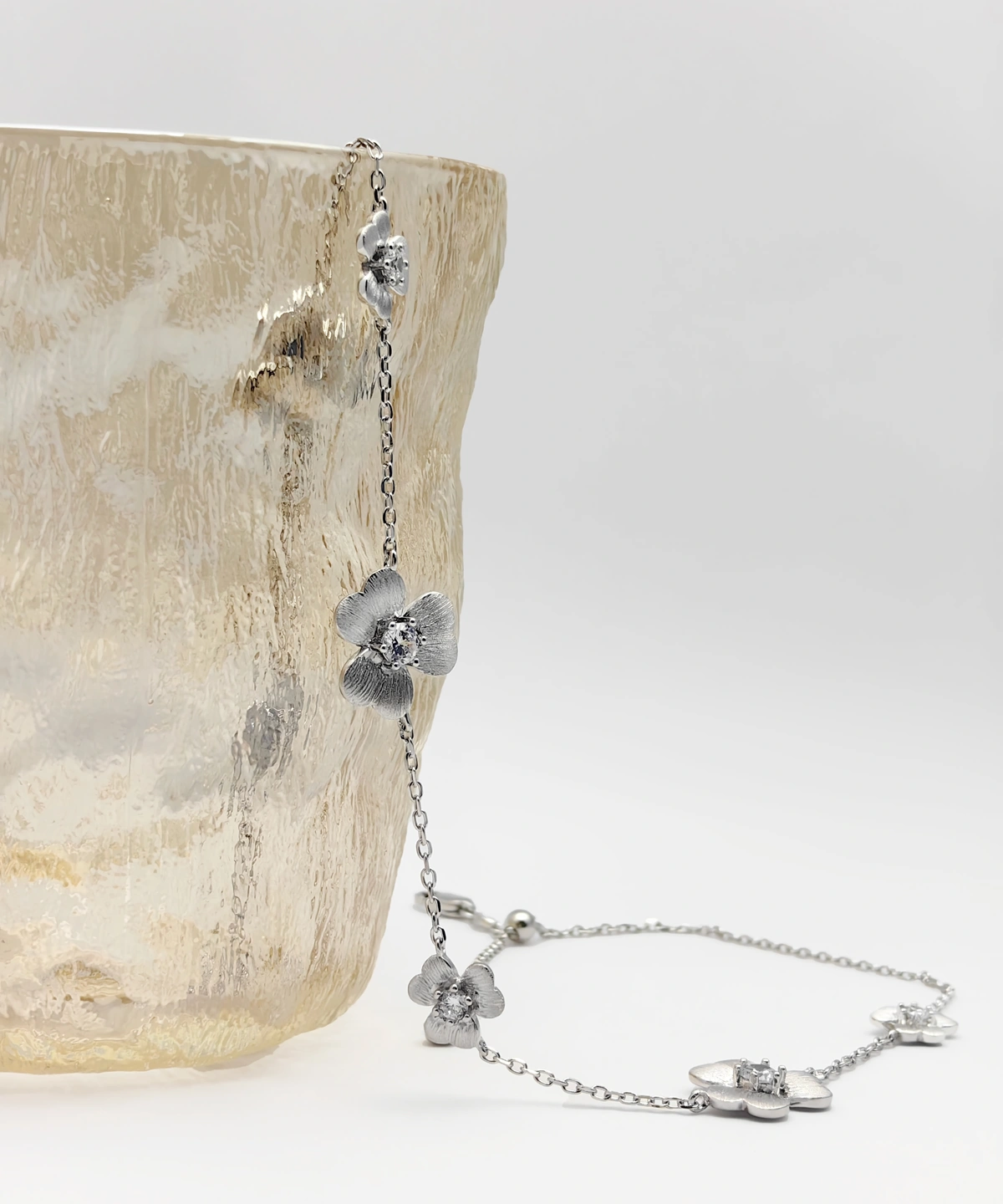 The image shows a delicate and elegant Sterling Silver Flower Necklace. The necklace is made of S925 Sterling Silver and features multiple flower-shaped pendants, each adorned with a sparkling 5A zirconia stone, resembling diamonds. The necklace is displayed draped over the edge of a textured, translucent glass vase, highlighting its intricate design and craftsmanship. The flowers are evenly spaced along the fine silver chain, showcasing the beauty and brilliance of the zirconia stones. This Diamond Flower Necklace is a stunning piece of jewelry that combines the timeless elegance of sterling silver with the dazzling sparkle of high-quality zirconia.