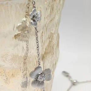 The image shows a delicate Sterling Silver Flower Necklace. This elegant piece is crafted from S925 Sterling Silver and features a flower-shaped pendant adorned with a sparkling 5A zirconia stone, resembling a diamond. The pendant has detailed petals with a brushed finish, adding to its realistic appearance. The necklace is draped over a textured, translucent vase, which highlights its intricate design and craftsmanship. The fine silver chain complements the pendant, enhancing the overall elegance of this diamond flower necklace.
