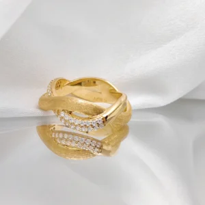 The image shows a beautifully crafted "Entwined Dimensions" ring. This intertwined ring is made of S925 sterling silver and plated with 18K gold. The design features two interwoven bands—one with a matte finish and the other adorned with sparkling 5A cubic zirconia stones. The ring is placed on a soft, white fabric, which enhances its elegance and highlights its intricate details. The perspective is slightly angled to showcase the intricate craftsmanship and the luxurious finish of the ring, making it the focal point of the image.
