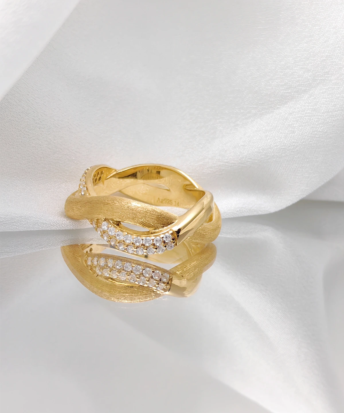 The image shows a beautifully crafted "Entwined Dimensions" ring. This intertwined ring is made of S925 sterling silver and plated with 18K gold. The design features two interwoven bands—one with a matte finish and the other adorned with sparkling 5A cubic zirconia stones. The ring is placed on a soft, white fabric, which enhances its elegance and highlights its intricate details. The perspective is slightly angled to showcase the intricate craftsmanship and the luxurious finish of the ring, making it the focal point of the image.