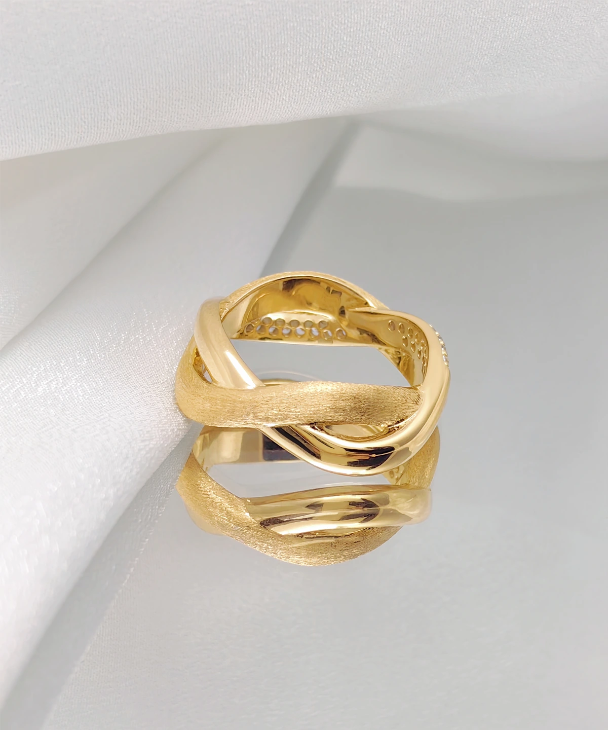 The image shows a beautifully crafted "Entwined Dimensions" ring. This intertwined ring is made of S925 sterling silver and is plated with 18K gold. It features two interwoven bands—one with a polished finish and the other with a matte finish. The ring is placed on a white surface, which highlights its intricate design and craftsmanship. The perspective showcases the elegant twist and the luxurious finish of the ring, making it the focal point of the image.