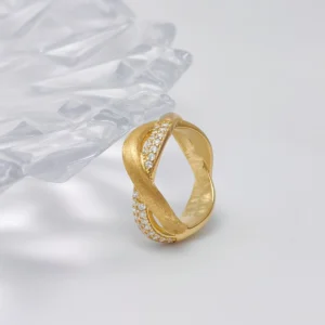 The image shows a beautifully crafted ring named "Entwined Dimensions." This intertwined ring is made of S925 sterling silver and is plated with 18K gold. The ring features a unique design where two bands twist around each other, with one band adorned with sparkling 5A cubic zirconia stones. The ring is placed on a white surface with a clear, faceted object in the background, adding an elegant touch. The perspective highlights the intricate details and the luxurious finish of the ring, making it the focal point.