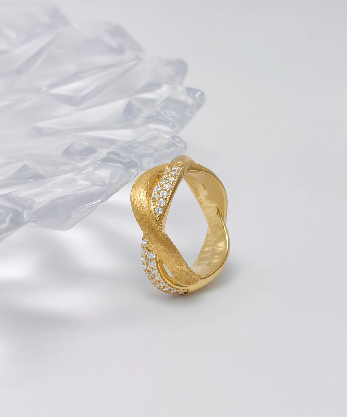 The image shows a beautifully crafted ring named "Entwined Dimensions." This intertwined ring is made of S925 sterling silver and is plated with 18K gold. The ring features a unique design where two bands twist around each other, with one band adorned with sparkling 5A cubic zirconia stones. The ring is placed on a white surface with a clear, faceted object in the background, adding an elegant touch. The perspective highlights the intricate details and the luxurious finish of the ring, making it the focal point.