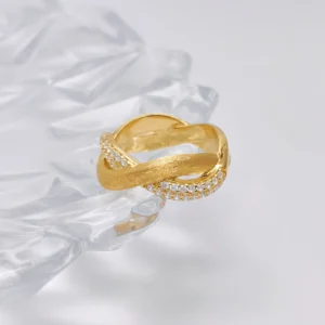 This image shows the "Entwined Dimensions" intertwined ring, which boasts an exquisite design. The ring is made of S925 sterling silver and is plated with 18K gold. The band features an intertwined design, combining a smooth, polished section with a section adorned with sparkling 5A cubic zirconia stones. The ring is placed on a clear, faceted surface, adding an elegant and sophisticated backdrop to the image. The perspective is slightly angled, highlighting the intricate details and elegant twist of the band.