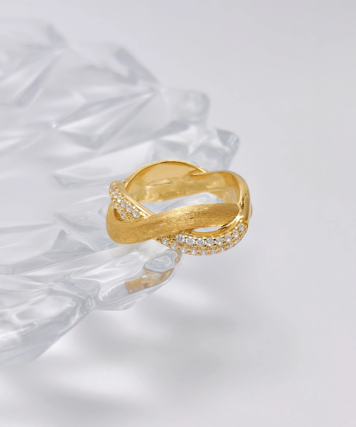 This image shows the "Entwined Dimensions" intertwined ring, which boasts an exquisite design. The ring is made of S925 sterling silver and is plated with 18K gold. The band features an intertwined design, combining a smooth, polished section with a section adorned with sparkling 5A cubic zirconia stones. The ring is placed on a clear, faceted surface, adding an elegant and sophisticated backdrop to the image. The perspective is slightly angled, highlighting the intricate details and elegant twist of the band.
