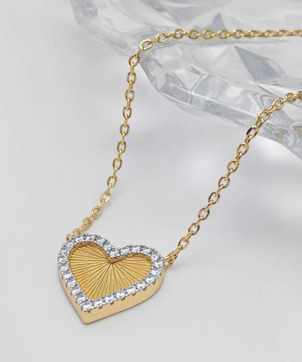 The image showcases a beautifully designed Gold Heart Pendant Necklace. This elegant piece is crafted from S925 Sterling Silver and plated with 18K gold. The heart-shaped pendant is adorned with sparkling 5A cubic zirconia stones along its edge, creating a dazzling border. The center of the pendant features a textured, sunburst pattern, adding a unique touch to the design. The pendant is suspended from a delicate gold link chain, also made from S925 Sterling Silver and plated with 18K gold. The necklace is displayed on a clear, faceted surface, which enhances its elegance and sophistication.