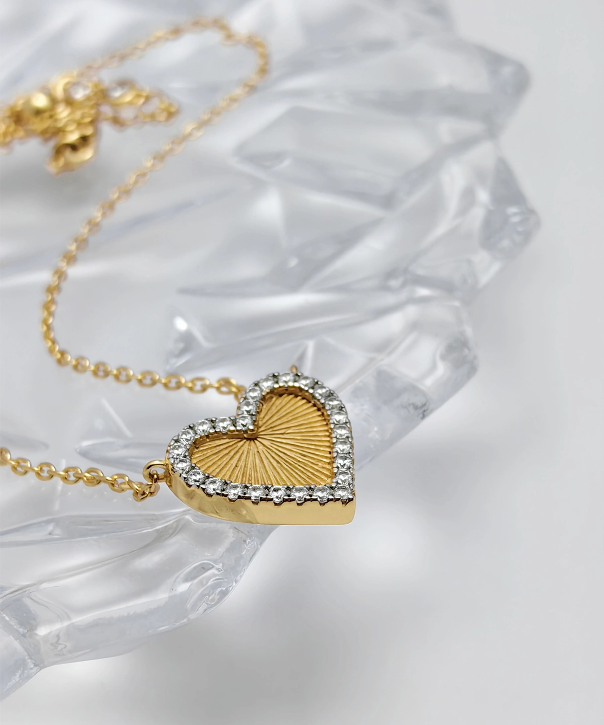 The image showcases a beautifully designed Gold Heart Pendant Necklace. This elegant piece is crafted from S925 Sterling Silver and plated with 18K gold. The heart-shaped pendant is adorned with sparkling 5A cubic zirconia stones along its edge, creating a dazzling border. The center of the pendant features a textured, sunburst pattern, adding a unique touch to the design. The pendant is suspended from a delicate gold link chain, also made from S925 Sterling Silver and plated with 18K gold. The necklace is displayed on a clear, faceted surface, which enhances its elegance and sophistication.