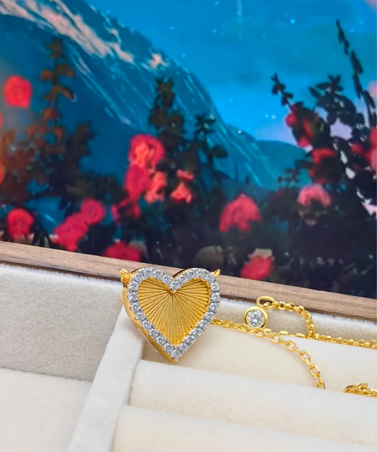 The image showcases a beautifully designed Gold Heart Pendant Necklace. This elegant piece is made from S925 Sterling Silver and plated with 18K gold. The pendant is heart-shaped and is adorned with 5A cubic zirconia stones around its edge, creating a sparkling border. The center of the heart has a textured sunburst design, adding depth and uniqueness. The pendant is suspended from a delicate gold chain, also made from S925 Sterling Silver and plated with 18K gold. The necklace is displayed in a cream-colored, velvet-lined jewelry box, enhancing its luxurious appearance. The background includes a scenic painting with mountains and flowers, adding a colorful and artistic touch to the presentation.