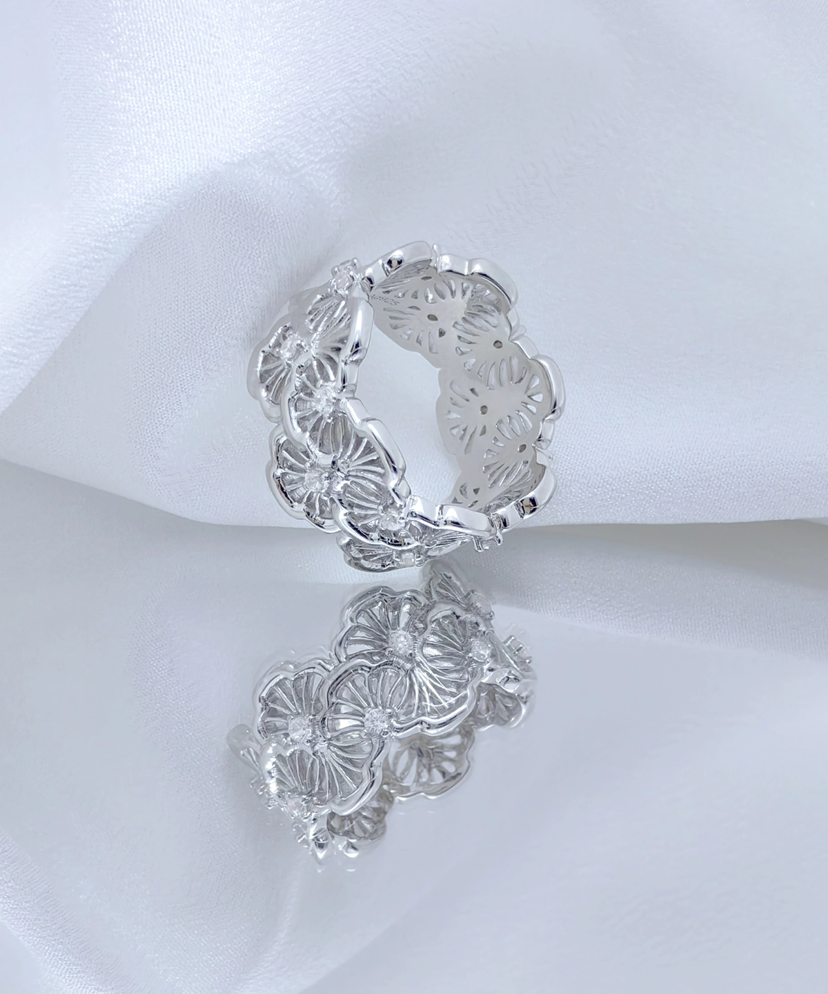 This image showcases a silver lace ring. The ring is made of rhodium-plated sterling silver and is set with 5A cubic zirconia stones. The design is very exquisite, with intricate floral pattern engravings on the surface of the ring, exhibiting an elegant and unique beauty. The floral patterns seem to encircle the entire ring, forming a continuous wreath. The ring is placed against a white fabric background, enhancing the ring's reflective effect and details. The ring's reflection in the mirror adds a visual element to the image. This design is not only artistic but also highlights the wearer's taste and status, making it very suitable for various formal occasions.