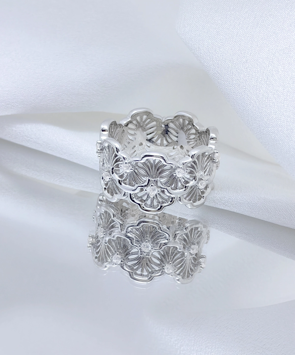 This image showcases a silver lace ring. The ring is made of rhodium-plated sterling silver and is set with 5A cubic zirconia stones. The design is very exquisite, with intricate floral pattern engravings on the surface of the ring, exhibiting an elegant and unique beauty. The floral patterns seem to encircle the entire ring, forming a continuous wreath. The ring is placed against a white fabric background, enhancing the ring's reflective effect and details. The ring's reflection in the mirror adds a visual element to the image. This design is not only artistic but also highlights the wearer's taste and status, making it very suitable for various formal occasions.
