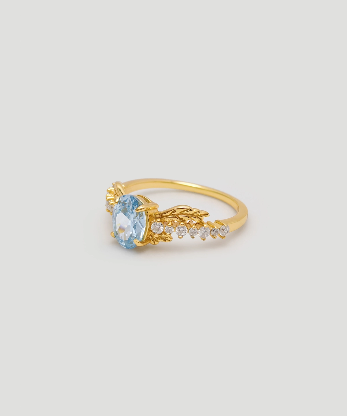 The image shows a Muse Goddess Gold Vermeil Lab Grown Sapphire Ring. This elegant ring features a central lab grown sapphire gemstone with a captivating blue hue, set within a gold vermeil band. The band is intricately designed with a series of small, sparkling 5A cubic zirconia stones that beautifully accentuate the main sapphire. Crafted from S925 sterling silver and coated with a layer of gold vermeil, the ring exudes luxury and sophistication. The ring is positioned at an angle, showcasing its detailed craftsmanship and the sparkle of the gemstones.