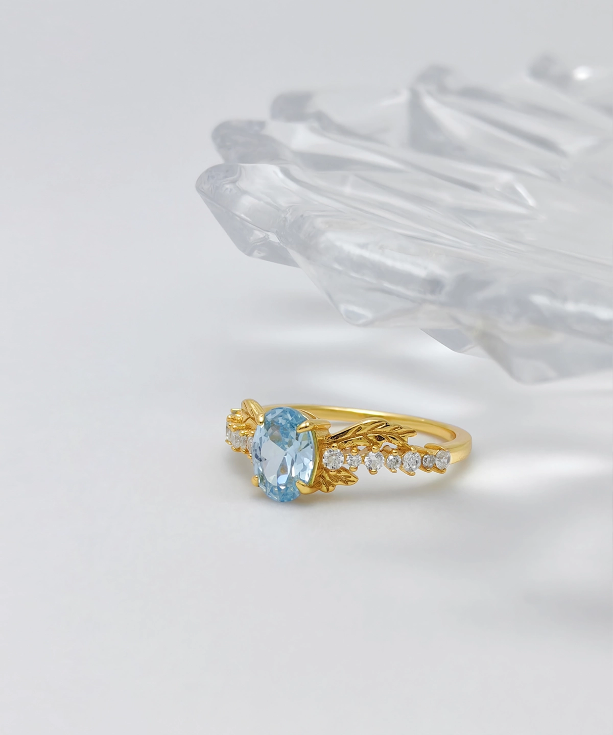 The image shows a Muse Goddess Gold Vermeil Lab Grown Sapphire Ring. This elegant ring features a large, oval-shaped light blue lab grown sapphire as the centerpiece. The gemstone is surrounded by a series of small, sparkling 5A cubic zirconia stones set along the intricately designed band, which resembles leaf-like patterns. Crafted from S925 sterling silver and coated with a layer of gold vermeil, the ring exudes luxury and sophistication. The ring is positioned front-facing on a white surface, with part of a clear, faceted object visible in the background. The combination of the blue gemstone, clear stones, and gold band makes the ring visually striking and elegant.