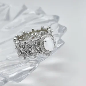 This image shows a beautifully crafted Sterling Silver Jade Ring. The ring is made of 925 sterling silver and is plated with rhodium for enhanced shine and durability. The central feature of the ring is a water foam jade, which is prominently set in the middle, surrounded by 5A cubic zirconia stones. The intricate design of the ring includes a pattern that resembles a crown, adding to its regal appearance. The ring is displayed on a clear, crystalline surface, which enhances its elegance and highlights the intricate details and sparkling stones.