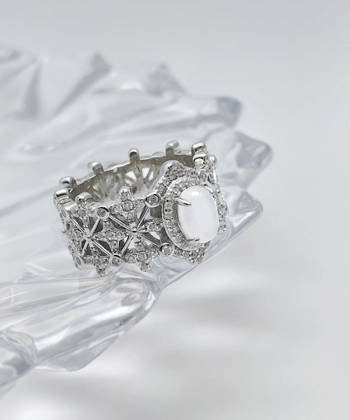 This image shows a beautifully crafted Sterling Silver Jade Ring. The ring is made of 925 sterling silver and is plated with rhodium for enhanced shine and durability. The central feature of the ring is a water foam jade, which is prominently set in the middle, surrounded by 5A cubic zirconia stones. The intricate design of the ring includes a pattern that resembles a crown, adding to its regal appearance. The ring is displayed on a clear, crystalline surface, which enhances its elegance and highlights the intricate details and sparkling stones.