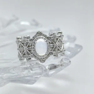 This image shows a beautifully crafted Sterling Silver Jade Ring. The ring is made of 925 sterling silver and is plated with rhodium for enhanced shine and durability. The central feature of the ring is a water foam jade, which is prominently set in the middle, surrounded by 5A cubic zirconia stones. The intricate design of the ring includes a pattern that resembles a crown, adding to its regal appearance. The ring is displayed on a clear, crystalline surface, which enhances its elegance and highlights the intricate details and sparkling stones.