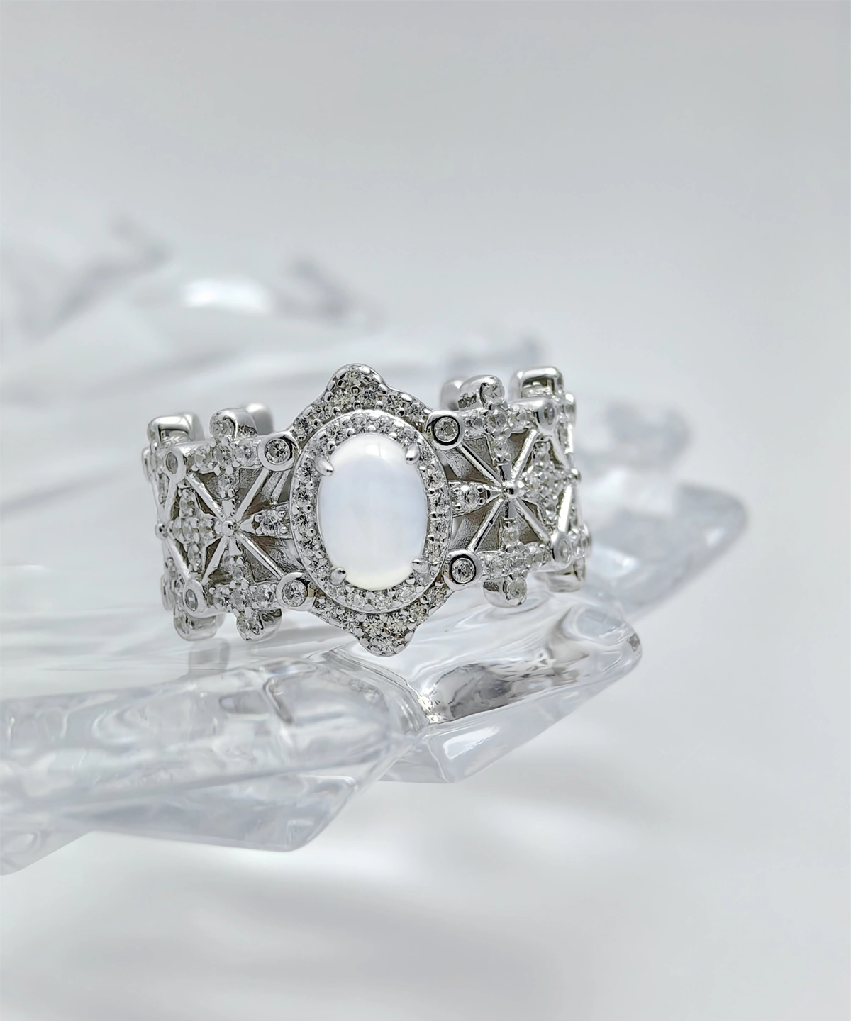 This image shows a beautifully crafted Sterling Silver Jade Ring. The ring is made of 925 sterling silver and is plated with rhodium for enhanced shine and durability. The central feature of the ring is a water foam jade, which is prominently set in the middle, surrounded by 5A cubic zirconia stones. The intricate design of the ring includes a pattern that resembles a crown, adding to its regal appearance. The ring is displayed on a clear, crystalline surface, which enhances its elegance and highlights the intricate details and sparkling stones.