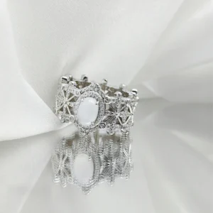 This image showcases a beautifully crafted Sterling Silver Jade Ring. The ring is made of S925 Sterling Silver plated with rhodium and is adorned with 5A cubic zirconia stones. The central feature of the ring is a water foam jade, which is an oval-shaped, translucent gemstone. The intricate design of the ring includes a lattice pattern with small cubic zirconia stones embedded throughout, giving it a regal and elegant appearance. The ring is displayed on a reflective mirror surface, which enhances its brilliance and detail. The background is a soft, white fabric that adds to its luxurious presentation.