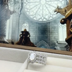 The image showcases a beautifully crafted Sterling Silver Jade Ring. This ring is made of 925 sterling silver and is plated with rhodium for enhanced shine and durability. It features an intricate design adorned with 5A cubic zirconia stones. The centerpiece of the ring is a water foam jade, an oval-shaped, translucent gemstone that adds elegance and sophistication to the piece. The ring is displayed in a luxurious jewelry box with a soft, cushioned interior, which highlights its elegance and craftsmanship. The background features an ornate, cathedral-like setting with gothic architectural elements, including a large circular stained glass window and detailed stone carvings. This elaborate setting adds to the ring's regal and sophisticated presentation.