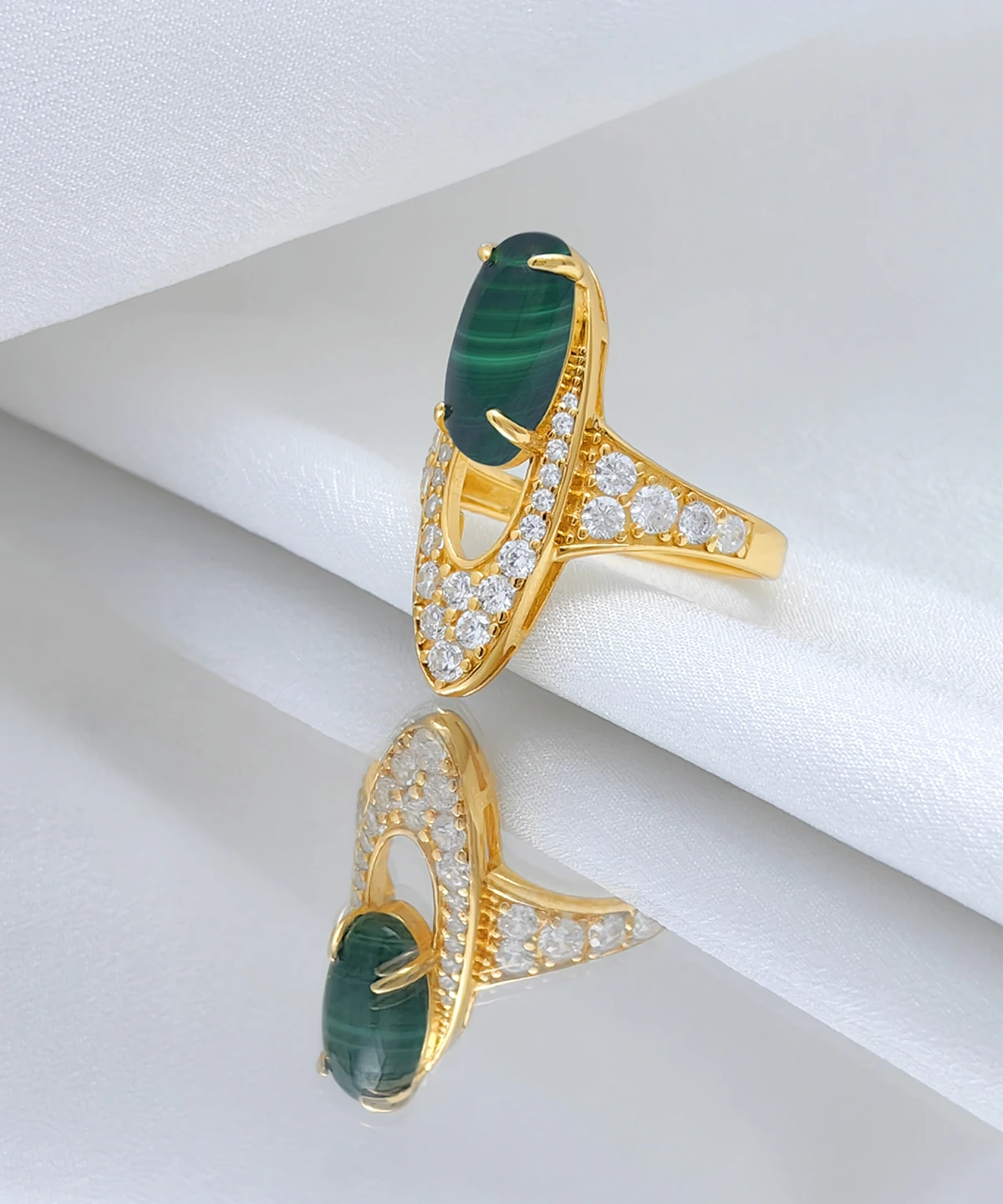 The image showcases a luxurious Gold Malachite Ring made from S925 Sterling Silver, plated with 18K gold. The ring features a prominent oval-shaped natural malachite stone set in the center, held by four prongs. Surrounding the malachite are numerous sparkling 5A cubic zirconia stones, intricately set into the gold-plated band, creating a dazzling effect. The ring is displayed elegantly on a reflective surface, with a white fabric backdrop that enhances its brilliance and detail. The reflection of the ring on the surface adds depth to the presentation, highlighting the craftsmanship and beauty of the piece. This is the Stardust of the Verdant Wilds Gold Vermeil Malachite Ring.