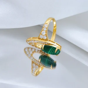 The image showcases a luxurious Gold Malachite Ring made from S925 Sterling Silver, plated with 18K gold. The ring features a prominent oval-shaped natural malachite stone set in the center, held by four prongs. Surrounding the malachite are numerous sparkling 5A cubic zirconia stones, intricately set into the gold-plated band, creating a dazzling effect. The ring is displayed elegantly on a reflective surface, with a white fabric backdrop that enhances its brilliance and detail. The reflection of the ring on the surface adds depth to the presentation, highlighting the craftsmanship and beauty of the piece. This is the Stardust of the Verdant Wilds Gold Vermeil Malachite Ring.