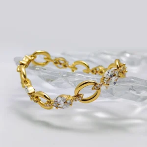 The image features a beautifully designed Gold Vermeil Diamond Link Bracelet. This elegant piece is crafted from S925 Sterling Silver and plated with 18K gold. The bracelet showcases a series of interlocking links, each adorned with clusters of sparkling 5A cubic zirconia stones. These gemstones are arranged in a combination of oval and round cuts, creating a dazzling effect. The bracelet is draped over a clear, textured surface, which enhances its luxurious and sophisticated appearance.