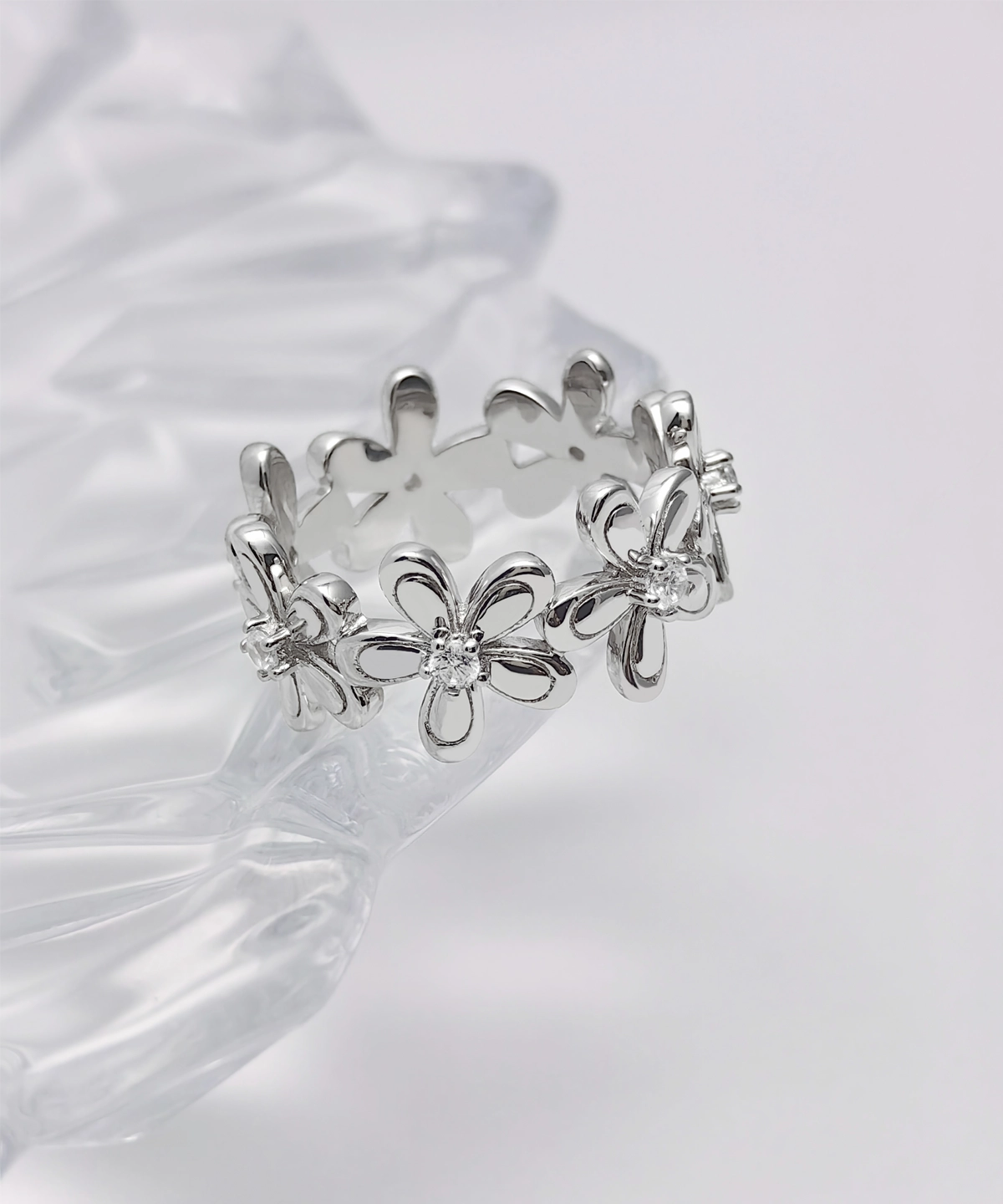 This image showcases the front view of the "Sunshine" silver flower ring, perfectly displaying the ring's overall design and details. The ring is standing upright on a transparent display stand, with a pure white background that highlights the exquisite design and high-quality materials of the ring. The main part of the ring is in the shape of flowers, made of sterling silver, with a smooth and shiny surface. Each flower's center is studded with 5A-grade zirconia, sparkling like morning dew. The arrangement of the flowers forms a circular pattern, making the ring look like a blooming floral wreath, exuding elegance and sophistication.