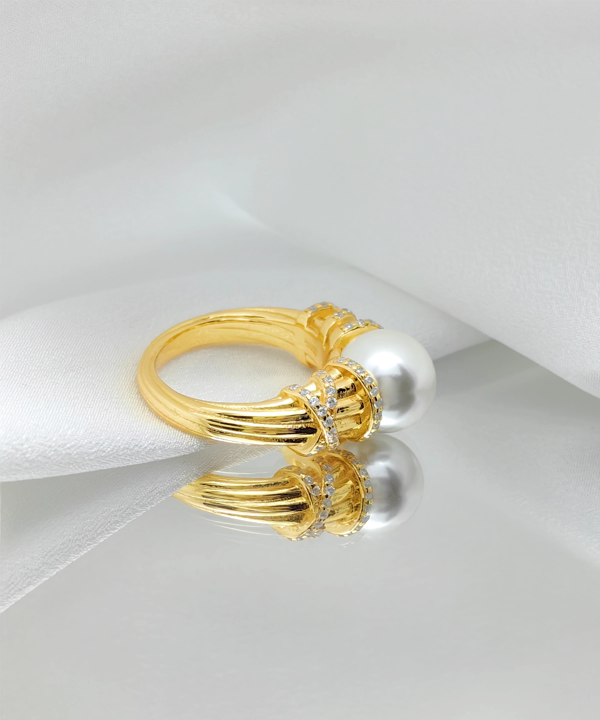 This image showcases a "Twilight" pearl ring. The ring is made of gold-plated on sterling silver, with multiple delicate textures on the surface of the band. At the top of the ring, there is a high-luster pearl, with a circle of small diamonds on each side, enhancing the ring's brilliance. The ring is placed on a piece of white fabric. The soft texture of the fabric contrasts sharply with the metallic sheen of the ring, highlighting the ring's exquisite and noble nature.