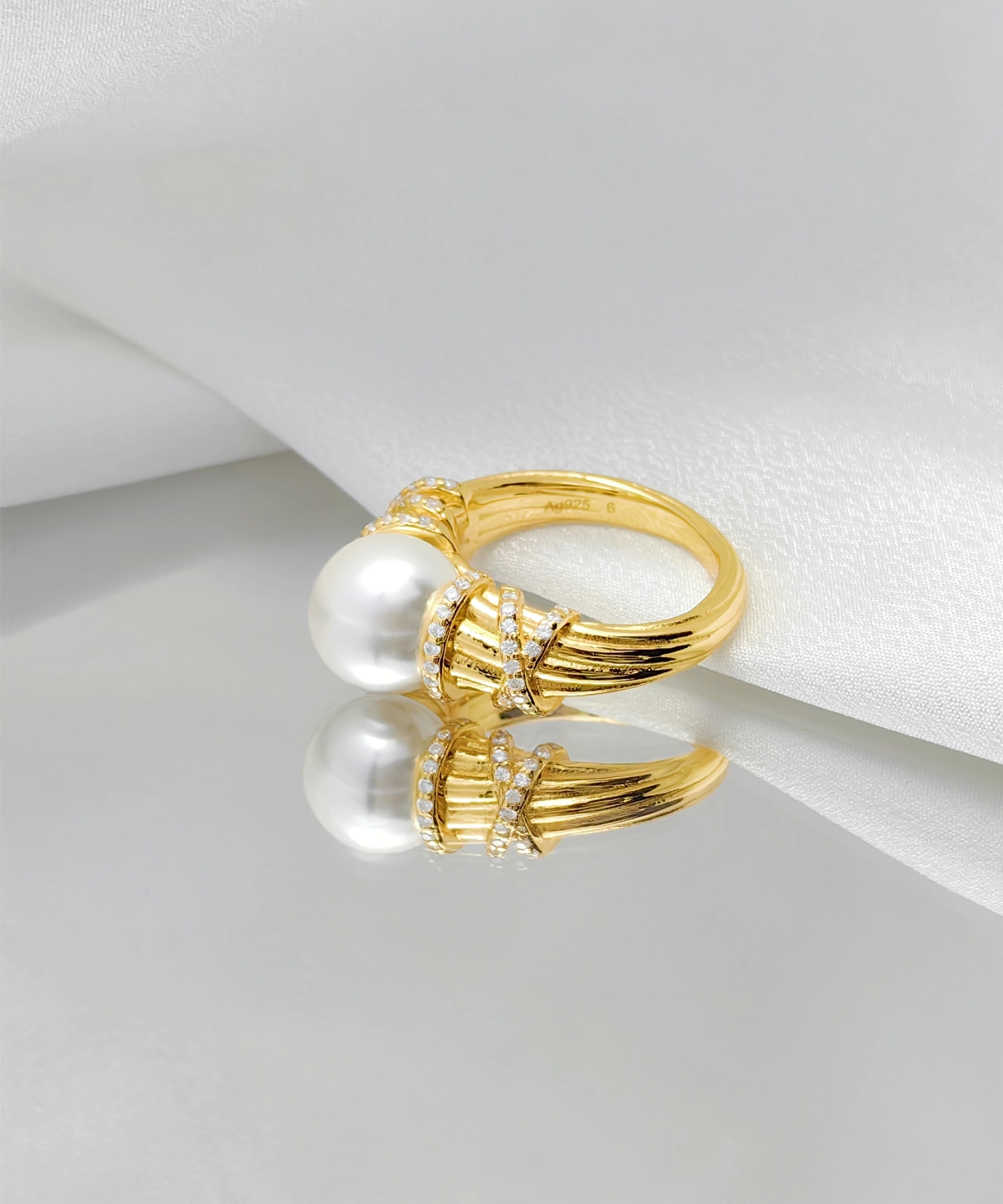 This image showcases a "Twilight" pearl ring. The ring is made of gold-plated on sterling silver, with multiple delicate textures on the surface of the band. At the top of the ring, there is a high-luster pearl, with a circle of small diamonds on each side, enhancing the ring's brilliance. The ring is placed on a piece of white fabric. The soft texture of the fabric contrasts sharply with the metallic sheen of the ring, highlighting the ring's exquisite and noble nature.