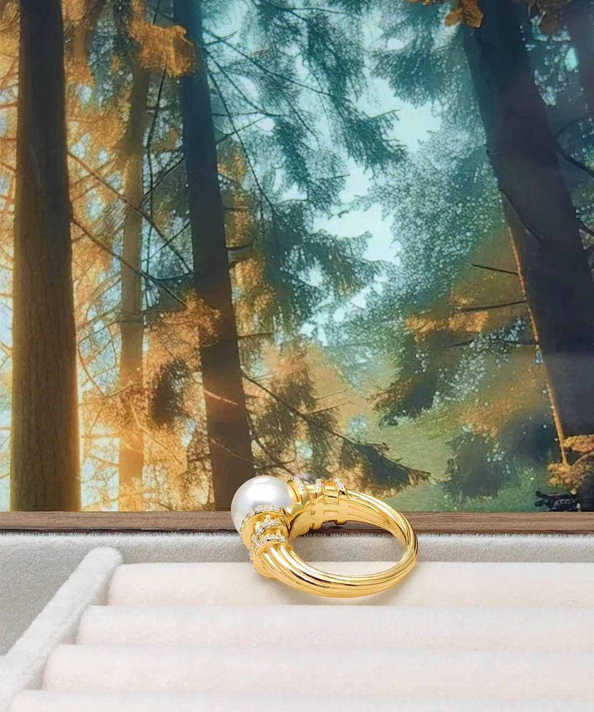 This image showcases a pearl ring named "Twilight." The ring is made of gold-plated on sterling silver, with a high-luster pearl embedded in the center of the ring face. The surface of the pearl is smooth, radiating a soft glow. The ring is placed in a soft ring display box, with a forest illustration in the background. Sunlight filters through the leaves, creating a tranquil and dreamy atmosphere. The design of the ring is exquisite, with delicate textures and decorations on the golden band. The pearl’s bright luster complements the overall elegant and noble appearance of the ring.