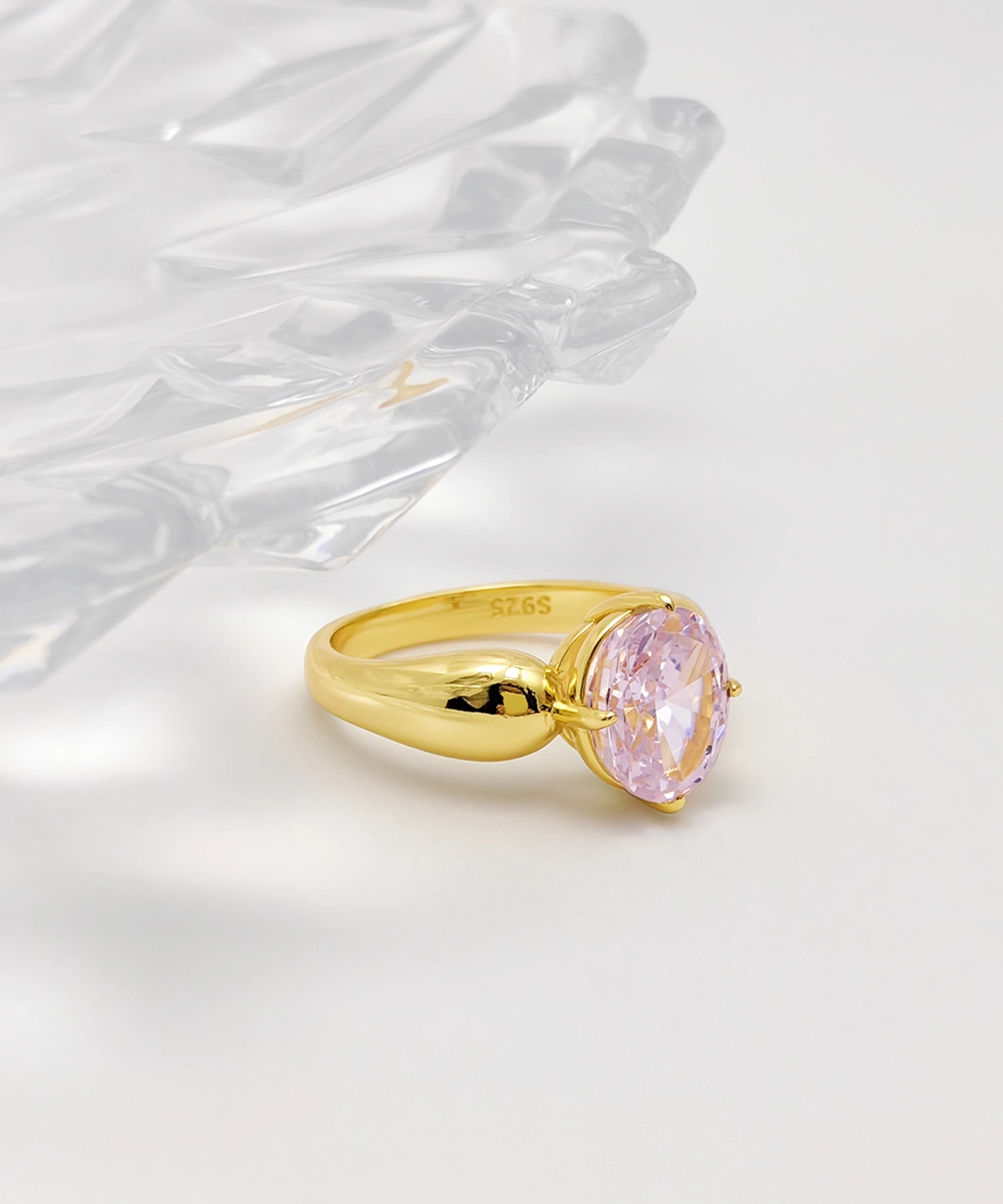 This image showcases an exquisite Lab Grown Pink Sapphire Ring. The ring features a band crafted from gold vermeil over sterling silver, and it is set with a lab-grown pink sapphire. The design of the ring is simple yet elegant, with the pink sapphire secured at the top by a four-prong setting. The ring is delicately displayed below a transparent glass plate, with soft lighting from behind that highlights the gold luster of the band and the vibrant color of the pink sapphire. This ring is not only a stylish accessory but also an ideal choice for women's jewelry collections.