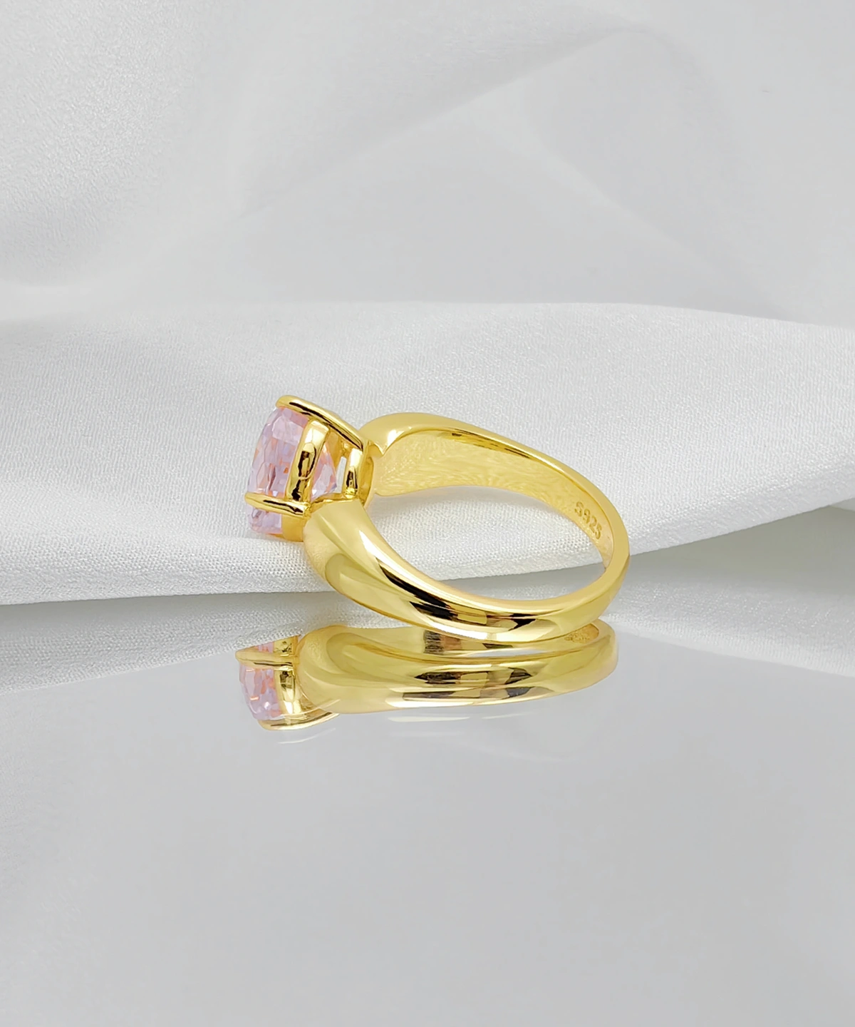 This image showcases a Lab Grown Pink Sapphire Ring. The ring features a band crafted from gold vermeil over sterling silver, set with a lab-grown pink sapphire. The ring is placed on a mirror and a piece of white fabric, which appears soft and lustrous. The design of the ring is simple yet elegant, with the gold band complementing the pink sapphire. The sapphire is secured at the top by a four-prong setting, giving it a delicate appearance. The inside of the ring is engraved with "925," indicating its sterling silver composition. The mirror's surface reflects the ring's luster, enhancing the overall beauty and display effect.