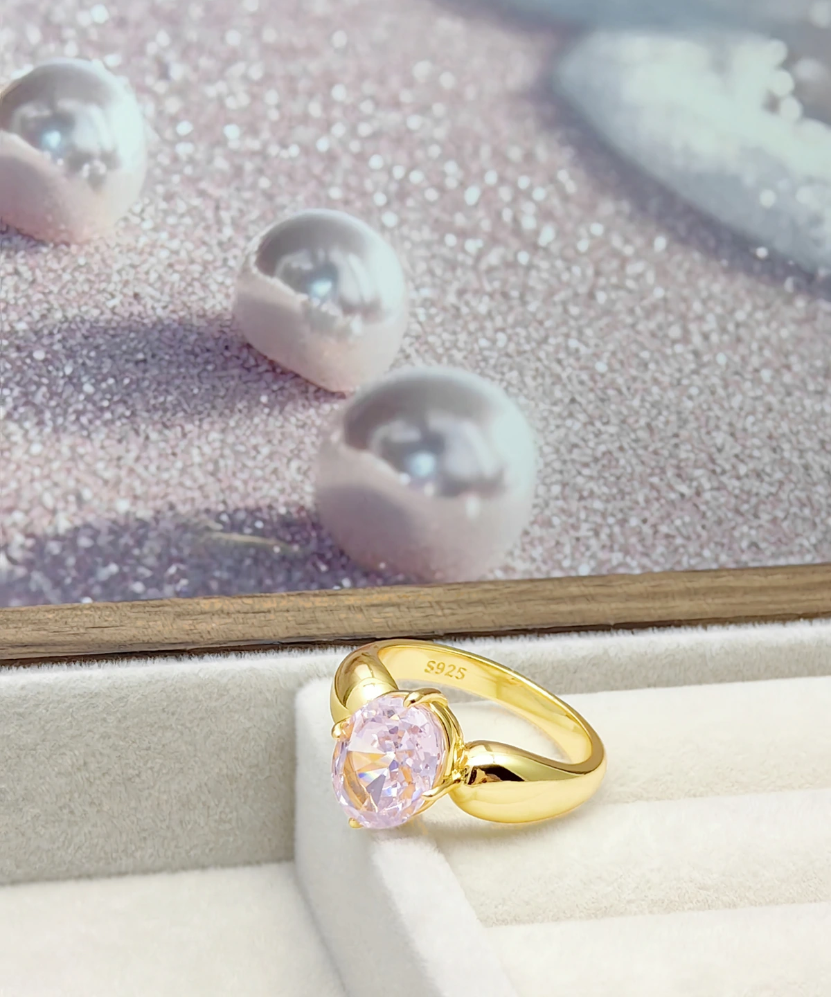 This image showcases a Lab Grown Pink Sapphire Ring. The ring features a band crafted from gold vermeil over sterling silver, set with a lab-grown pink sapphire. The ring is elegantly placed in a soft jewelry display box, with a background of scattered smooth pink pearls. The design of the ring is simple yet elegant, with the gold band complementing the pink sapphire. The inside of the ring is engraved with "S925," indicating its sterling silver composition.