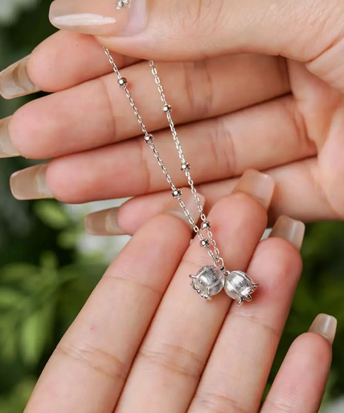 The image presents a close-up of a delicate bracelet. Crafted from sterling silver, the bracelet features evenly spaced small beads along its chain. The focal point of the piece is a pair of lily of the valley-inspired charms, meticulously designed to capture the natural elegance of the flowers. Two well-manicured hands gently hold the bracelet, emphasizing its exquisite craftsmanship and graceful design. The slightly blurred background, with hints of greenery, enhances the bracelet’s prominence, adding a touch of natural beauty to the scene.