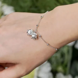 The image is a close-up of a wrist adorned with a sterling silver lily of the valley bracelet. Crafted from sterling silver, this delicate piece features a fine chain with evenly spaced small beads. Two intricately designed lily of the valley charms dangle gracefully from the chain, adding a touch of elegance and charm. The hand wearing the bracelet is set against a softly blurred background, highlighting the bracelet’s refined and graceful design.