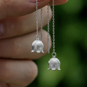 The image shows a close-up of a hand holding a pair of Sterling Silver Lily Of The Valley Earrings. Each earring features a delicate, elongated design that resembles the lily of the valley flower. The earrings are intricately detailed with small, bell-shaped blooms and finely textured petals, crafted from sterling silver. The earrings dangle gracefully from a thin, silver chain, adding to their elegant appearance. The background is softly blurred, highlighting the intricate craftsmanship and shiny finish of the earrings. These Sterling Silver Lily Of The Valley Earrings are beautiful and refined, making them a charming accessory for any outfit.