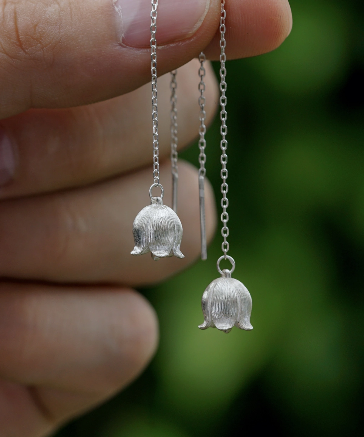 The image shows a close-up of a hand holding a pair of Sterling Silver Lily Of The Valley Earrings. Each earring features a delicate, elongated design that resembles the lily of the valley flower. The earrings are intricately detailed with small, bell-shaped blooms and finely textured petals, crafted from sterling silver. The earrings dangle gracefully from a thin, silver chain, adding to their elegant appearance. The background is softly blurred, highlighting the intricate craftsmanship and shiny finish of the earrings. These Sterling Silver Lily Of The Valley Earrings are beautiful and refined, making them a charming accessory for any outfit.