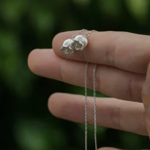 The image shows a close-up of a hand holding a pair of Sterling Silver Lily Of The Valley Earrings. Each earring features a delicate, elongated design that resembles the lily of the valley flower. The earrings are intricately detailed with small, bell-shaped blooms and finely textured petals, crafted from sterling silver. The earrings dangle gracefully from a thin, silver chain, adding to their elegant appearance. The background is softly blurred, highlighting the intricate craftsmanship and shiny finish of the earrings. These Sterling Silver Lily Of The Valley Earrings are beautiful and refined, making them a charming accessory for any outfit.