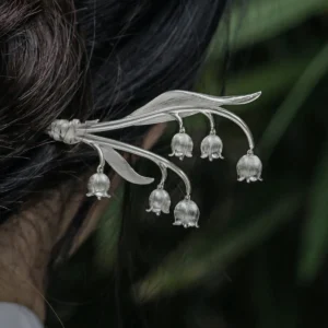The image is a close-up shot from behind, capturing a model with her hair styled in a bun, adorned with a sterling silver lily of the valley hairpin. The intricately designed hairpin features gracefully curved stems with multiple delicate bell-shaped blossoms, resembling the natural beauty of the lily of the valley. The model wears a light-colored garment with an elegant floral pattern. The softly blurred background of green leaves contrasts with the silver hair accessory, making it stand out. This photograph beautifully highlights the hairpin’s refined and graceful design, showcasing its craftsmanship and aesthetic appeal.