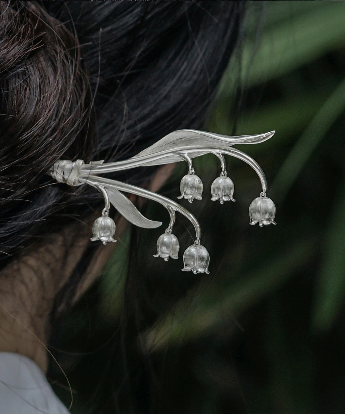 The image is a close-up shot from behind, capturing a model with her hair styled in a bun, adorned with a sterling silver lily of the valley hairpin. The intricately designed hairpin features gracefully curved stems with multiple delicate bell-shaped blossoms, resembling the natural beauty of the lily of the valley. The model wears a light-colored garment with an elegant floral pattern. The softly blurred background of green leaves contrasts with the silver hair accessory, making it stand out. This photograph beautifully highlights the hairpin’s refined and graceful design, showcasing its craftsmanship and aesthetic appeal.