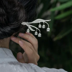 The image is a close-up shot from behind, capturing a model with her hair styled in a bun, adorned with a sterling silver lily of the valley hairpin. The intricately designed hairpin features gracefully curved stems with multiple delicate bell-shaped blossoms, resembling the natural beauty of the lily of the valley. The model wears a light-colored garment with an elegant floral pattern. The softly blurred background of green leaves contrasts with the silver hair accessory, making it stand out. This photograph beautifully highlights the hairpin’s refined and graceful design, showcasing its craftsmanship and aesthetic appeal.