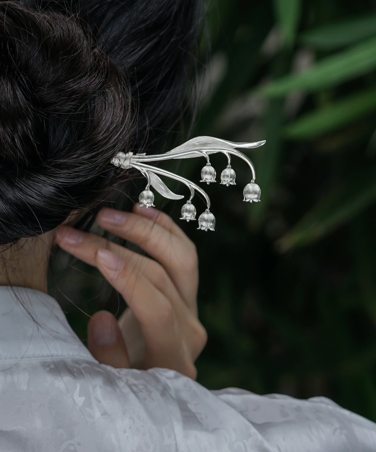 The image is a close-up shot from behind, capturing a model with her hair styled in a bun, adorned with a sterling silver lily of the valley hairpin. The intricately designed hairpin features gracefully curved stems with multiple delicate bell-shaped blossoms, resembling the natural beauty of the lily of the valley. The model wears a light-colored garment with an elegant floral pattern. The softly blurred background of green leaves contrasts with the silver hair accessory, making it stand out. This photograph beautifully highlights the hairpin’s refined and graceful design, showcasing its craftsmanship and aesthetic appeal.