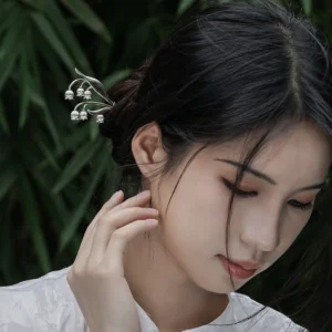The image is a front-facing close-up of a model wearing a sterling silver lily of the valley hairpin. The hairpin features an intricate design with delicate silver stems and small bell-shaped blossoms, mimicking the natural elegance of the lily of the valley plant. It is gracefully placed in the model’s dark hair, which is styled into an elegant bun. The model wears a high-collared white garment made of subtly lustrous fabric. The softly blurred background reveals hints of green foliage, adding a natural touch. This image highlights the fine craftsmanship and elegance of the sterling silver lily of the valley hairpin, showcasing its aesthetic appeal and how it can be worn as a decorative accessory.