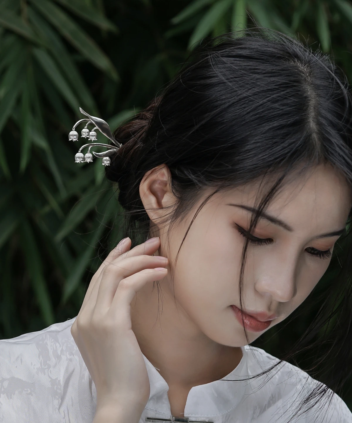 The image is a front-facing close-up of a model wearing a sterling silver lily of the valley hairpin. The hairpin features an intricate design with delicate silver stems and small bell-shaped blossoms, mimicking the natural elegance of the lily of the valley plant. It is gracefully placed in the model’s dark hair, which is styled into an elegant bun. The model wears a high-collared white garment made of subtly lustrous fabric. The softly blurred background reveals hints of green foliage, adding a natural touch. This image highlights the fine craftsmanship and elegance of the sterling silver lily of the valley hairpin, showcasing its aesthetic appeal and how it can be worn as a decorative accessory.