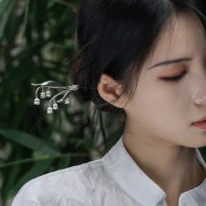 The image is a front-facing close-up of a model wearing a sterling silver lily of the valley hairpin. The hairpin features an intricate design with delicate silver stems and small bell-shaped blossoms, mimicking the natural elegance of the lily of the valley plant. It is gracefully placed in the model’s dark hair, which is styled into an elegant bun. The model wears a high-collared white garment made of subtly lustrous fabric. The softly blurred background reveals hints of green foliage, adding a natural touch. This image highlights the fine craftsmanship and elegance of the sterling silver lily of the valley hairpin, showcasing its aesthetic appeal and how it can be worn as a decorative accessory.