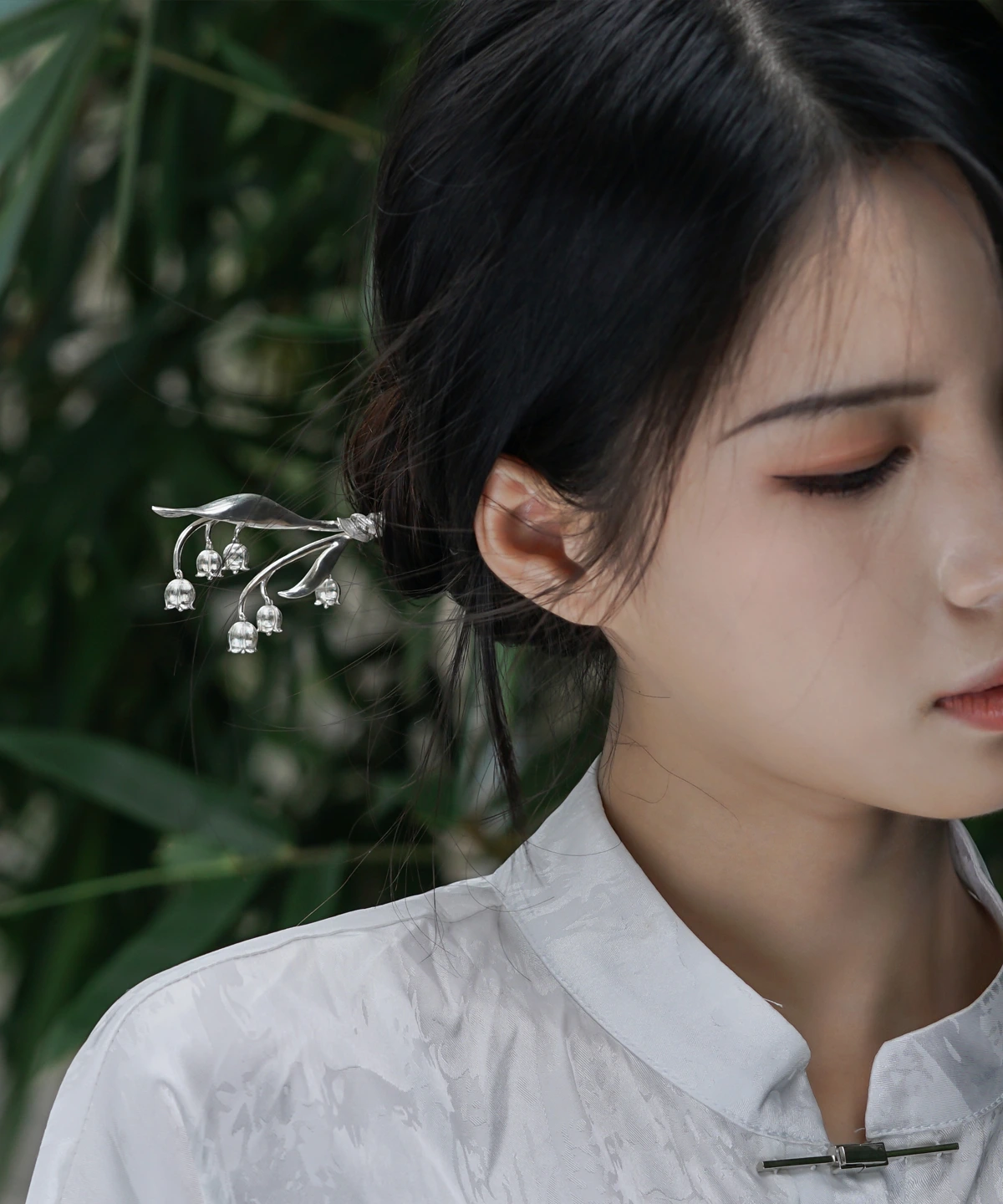 The image is a front-facing close-up of a model wearing a sterling silver lily of the valley hairpin. The hairpin features an intricate design with delicate silver stems and small bell-shaped blossoms, mimicking the natural elegance of the lily of the valley plant. It is gracefully placed in the model’s dark hair, which is styled into an elegant bun. The model wears a high-collared white garment made of subtly lustrous fabric. The softly blurred background reveals hints of green foliage, adding a natural touch. This image highlights the fine craftsmanship and elegance of the sterling silver lily of the valley hairpin, showcasing its aesthetic appeal and how it can be worn as a decorative accessory.