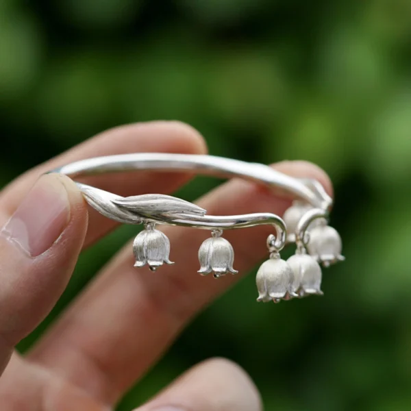 The image showcases a sterling silver open bangle inspired by lily of the valley. Crafted from sterling silver, this open-design bangle features delicate details, with several finely crafted bell-shaped flowers gracefully hanging from it. The meticulous craftsmanship of these blossoms adds a touch of elegance to the piece. The bangle itself has a smooth, polished surface that enhances its refined appearance. The softly blurred background highlights the intricate details of the bracelet, making it stand out. This sterling silver lily of the valley open bangle is a beautiful and elegant accessory that embodies a nature-inspired design.