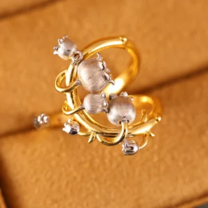 The image shows a beautifully crafted ring named Ethereal Bells | Sterling Silver Lily Of The Valley Ring. The ring features a design inspired by the lily of the valley flower, with delicate, bell-shaped flowers made of sterling silver. These flowers are attached to a gold-plated band that has an intricate, vine-like structure. The combination of the silver flowers and the gold-plated band creates a striking contrast, highlighting the detailed craftsmanship and elegance of the piece. The ring is set against a soft, beige background, which enhances the visual appeal of the jewelry. This Ethereal Bells | Sterling Silver Lily Of The Valley Ring is a stunning example of artistic design and fine materials, making it a desirable addition to any jewelry collection.
