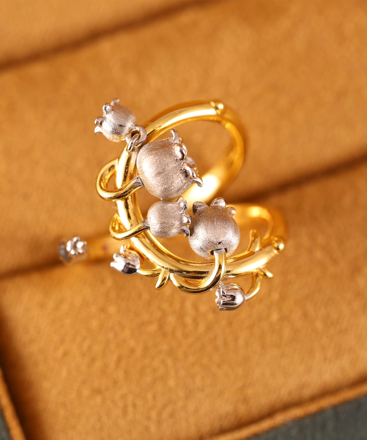 The image shows a beautifully crafted ring named Ethereal Bells | Sterling Silver Lily Of The Valley Ring. The ring features a design inspired by the lily of the valley flower, with delicate, bell-shaped flowers made of sterling silver. These flowers are attached to a gold-plated band that has an intricate, vine-like structure. The combination of the silver flowers and the gold-plated band creates a striking contrast, highlighting the detailed craftsmanship and elegance of the piece. The ring is set against a soft, beige background, which enhances the visual appeal of the jewelry. This Ethereal Bells | Sterling Silver Lily Of The Valley Ring is a stunning example of artistic design and fine materials, making it a desirable addition to any jewelry collection.